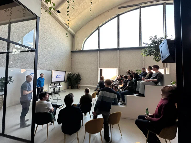 Bezoekers op stoelen en een tribune luisteren in een lichte zaal met boogramen